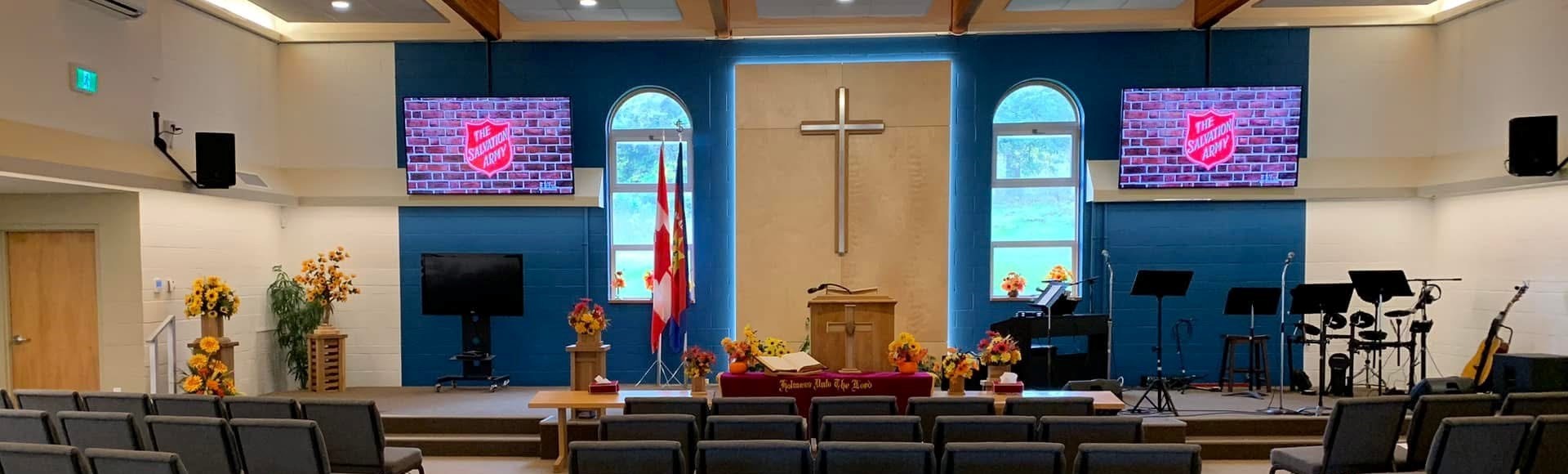 Interior of the Nanaimo Sanctuary with rows of seating facing a central altar, featuring a wooden pulpit beneath a large cross, arched windows, floral arrangements, flags, and a worship stage with musical instruments and screens.